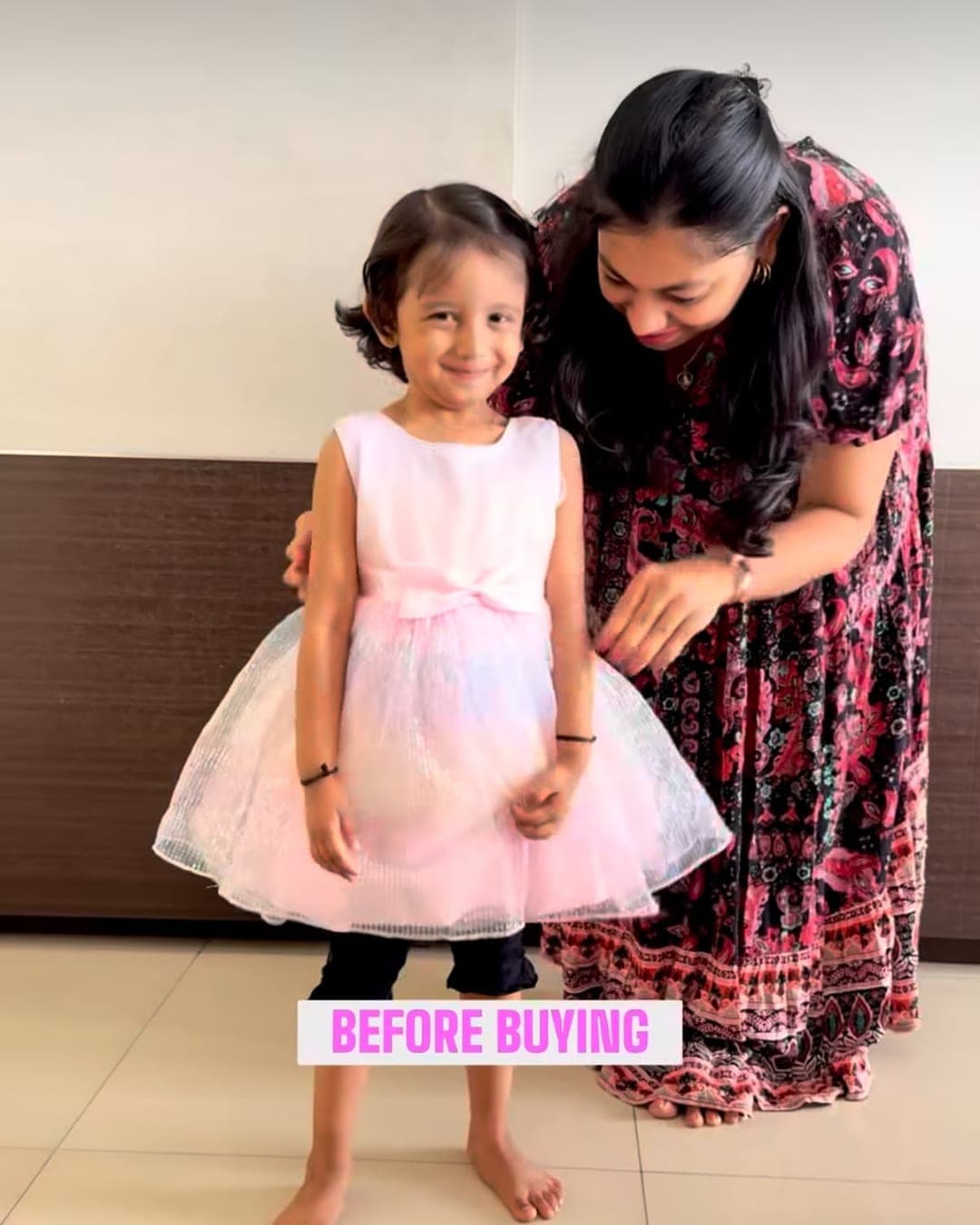 A young girl in a pink party dress standing at home while her mother adjusts the hem, during a Skipzy home try-on