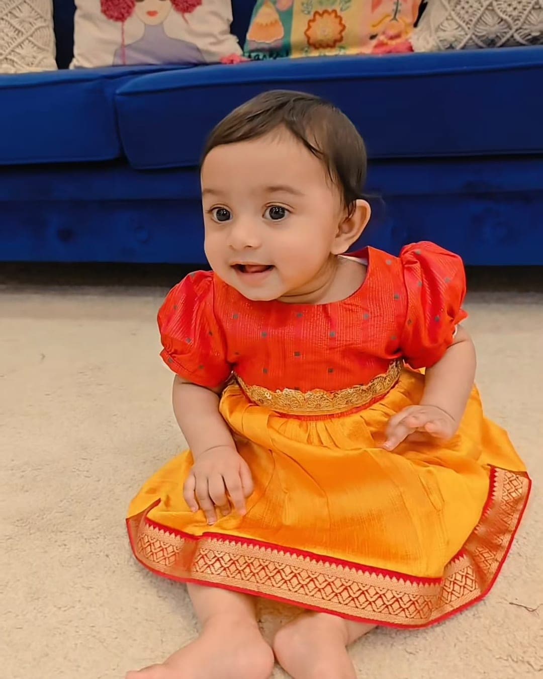A baby girl sitting on the floor in an orange and red ethnic dress tried on at home