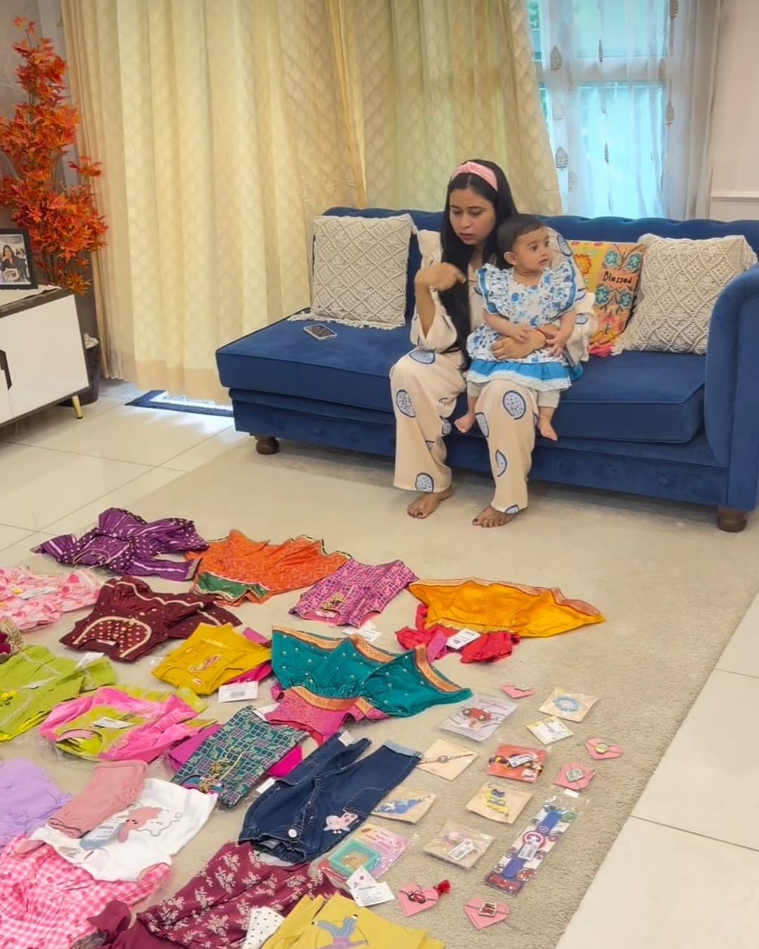 A mother sitting on a sofa holding her baby, with dozens of kids outfits laid out across the floor in front of them