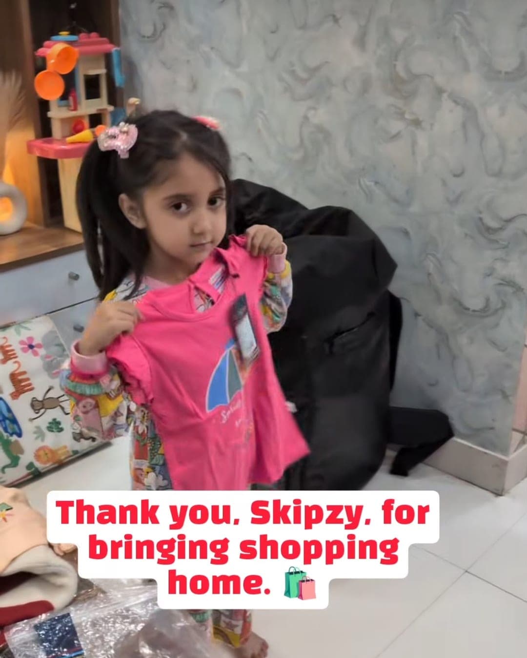A girl holding up a pink top on a hanger against herself at home, with more outfits spread out around her